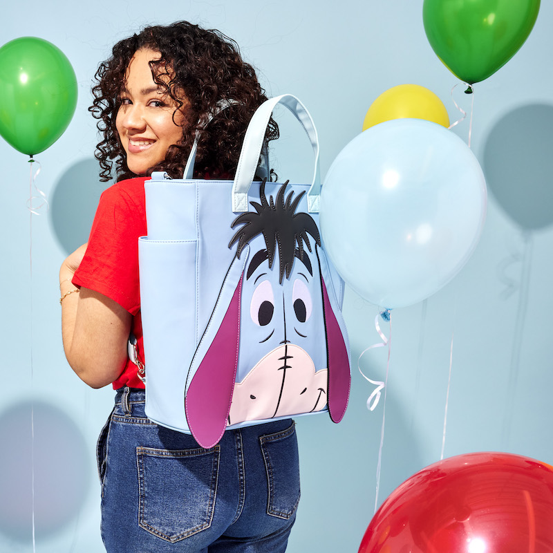 Image of woman standing against a blue background with balloons with her back to camera wearing the Loungefly Winnie the Pooh Eeyore Convertible Backpack and Tote Bag and smiling back at the camera 